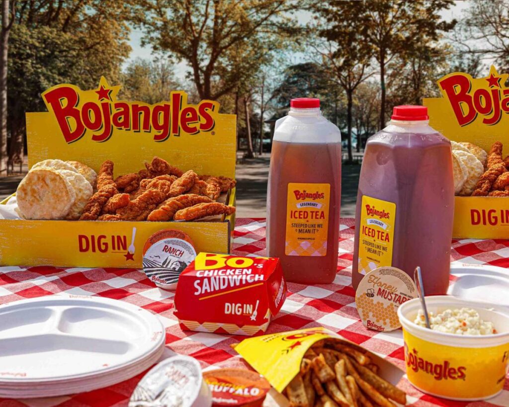 Chicken fingers, biscuits, French fires, cole slaw and plastic jugs of iced tea from Bojangles displayed on a picnic table with a red checked tablecloth