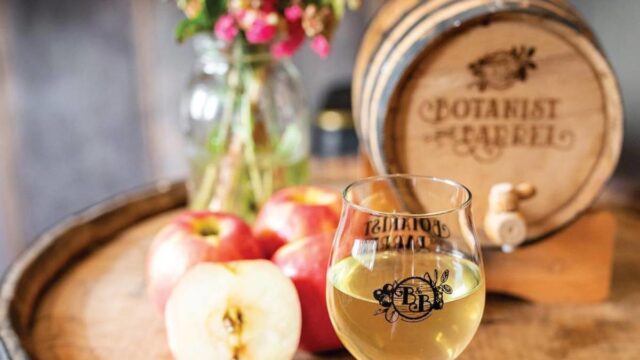 A glass of cider presented on top of a barrel with apples and colorful flowers