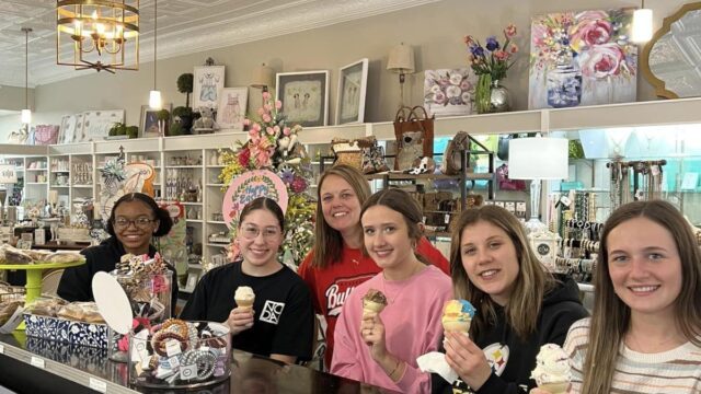 Five young women sit at a counter holding ice cream cones and smiling with an adult woman smiling and standing behind them