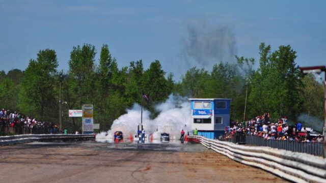 Two motor cars emitting plumes of white smoke at the start of a race
