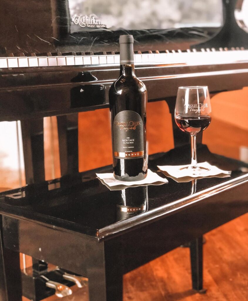 A bottle of wine and a partially full glass of wine sit on white square napkins, on a piano bench in front of a piano